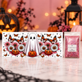A Halloween-themed decorative card or box featuring a white ghost, pumpkins, flowers, and orange butterflies, displayed next to a small clear bag filled with pink glitter on a polished marble surface. The background is softly blurred with warm lights and Halloween decor.