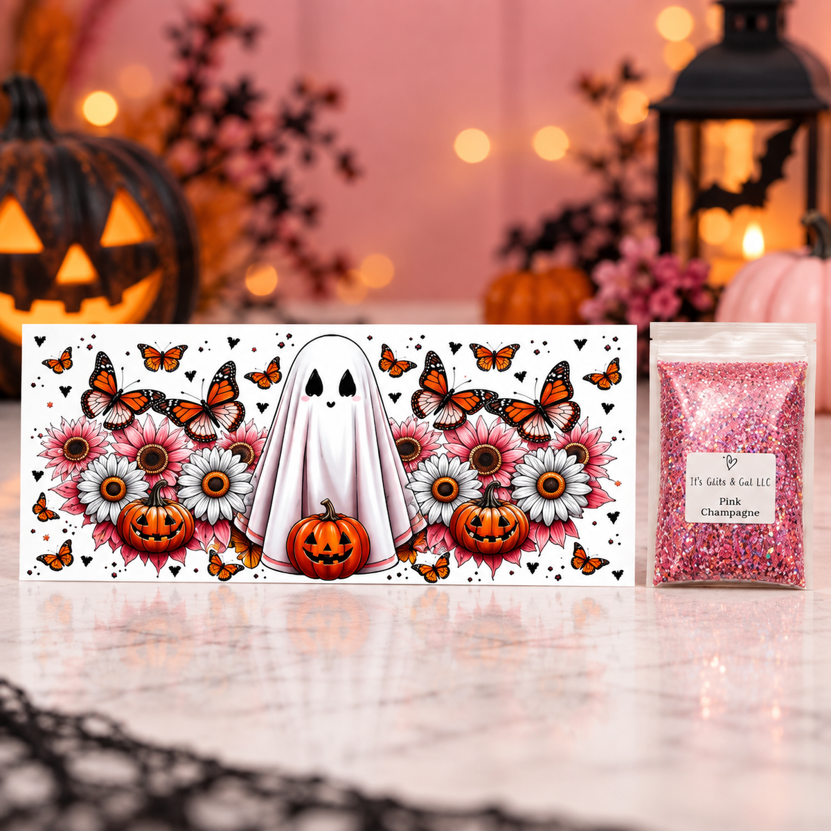 A Halloween-themed decorative card or box featuring a white ghost, pumpkins, flowers, and orange butterflies, displayed next to a small clear bag filled with pink glitter on a polished marble surface. The background is softly blurred with warm lights and Halloween decor.
